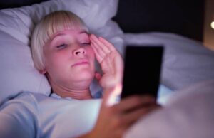 Junge Frau liegt nachts im Bett und schaut ins Handy – Blaulicht stört das Schlafhormon Melatonin