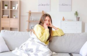 Frau sitzt müde auf dem Sofa unter einer Decke – Symbolbild für Müdigkeit im Winter